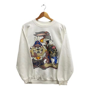 Op de afbeelding: Witte trui met een grafische print van Bugs Bunny en Taz van Looney Tunes. Bugs Bunny draagt een zwart leren jack en Taz draagt een geruit overhemd en een baseballpet.