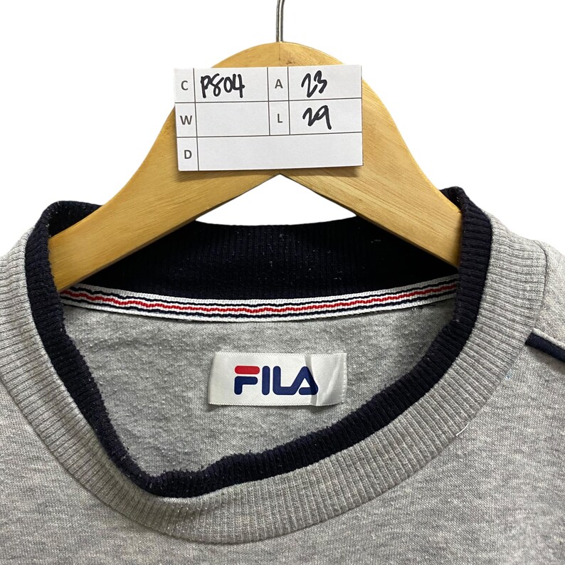 Puede incluir: Sudadera FILA gris con cuello y ribetes azul marino. La sudadera tiene una etiqueta blanca con el logo de FILA en rojo y azul. La prenda est&aacute; colgada de una percha de madera.
