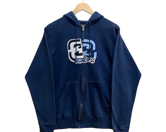 Può includere: Felpa blu navy con zip e tasca frontale. Il logo "Billabong" bianco, azzurro e nero è sul petto. La felpa è appesa a una gruccia di legno.
