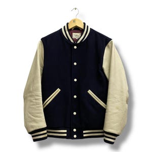 May include: A navy blue varsity jacket with cream-colored leather sleeves. The jacket features white and black striped cuffs and collar, white buttons, and two front pockets. The jacket is hanging on a wooden hanger.