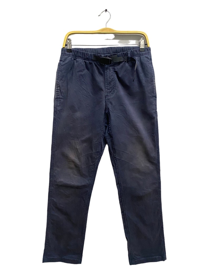 May include: Navy blue pants featuring an elastic waistband with an integrated black nylon webbing belt and a plastic buckle closure. The pants have a straight leg cut, side pockets, and visible stitching details along the seams and leg panels.