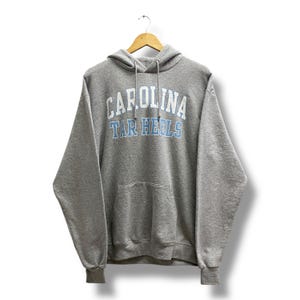 May include: A heather gray hooded sweatshirt with the text "CAROLINA TAR HEELS" in white and light blue. The sweatshirt has a front pocket and a drawstring hood. The garment is hanging on a wooden hanger.