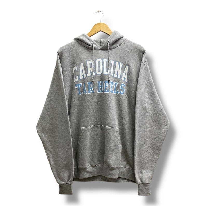 May include: A heather gray hooded sweatshirt with the text "CAROLINA TAR HEELS" in white and light blue. The sweatshirt has a front pocket and a drawstring hood. The garment is hanging on a wooden hanger.