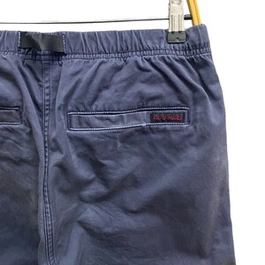 May include: Navy blue pants featuring an elastic waistband with an integrated black nylon belt. The back includes a single welt pocket with a small rectangular label displaying the red embroidered text Gramicci, all hanging from a wooden hanger.