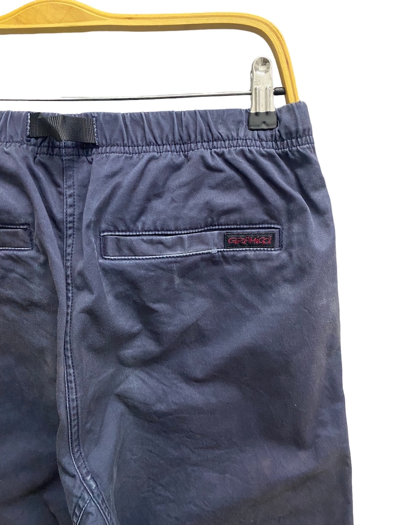 May include: Navy blue pants featuring an elastic waistband with an integrated black nylon belt. The back includes a single welt pocket with a small rectangular label displaying the red embroidered text Gramicci, all hanging from a wooden hanger.