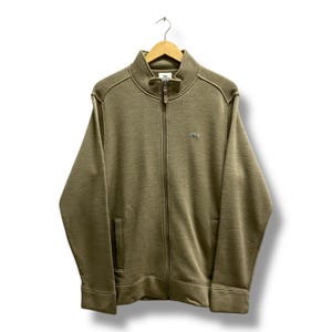 May include: A full-zip olive green jacket with a high collar and two front pockets. The jacket features the Lacoste logo on the chest and is hanging on a wooden hanger.