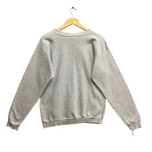 May include: A light gray long-sleeve crewneck sweatshirt made of a heathered fabric. The garment features a ribbed collar, ribbed cuffs, and a ribbed hemline. It is displayed hanging on a wooden hanger against a plain white background.