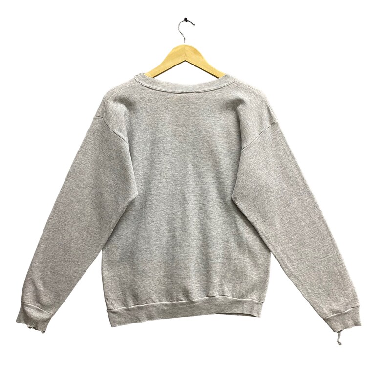 May include: A light gray long-sleeve crewneck sweatshirt made of a heathered fabric. The garment features a ribbed collar, ribbed cuffs, and a ribbed hemline. It is displayed hanging on a wooden hanger against a plain white background.