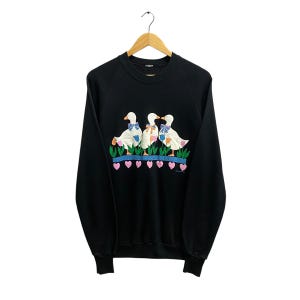 May include: Black sweatshirt with a graphic of three white ducks wearing blue bows and standing on a green patch with pink hearts. The ducks are surrounded by green leaves and flowers.