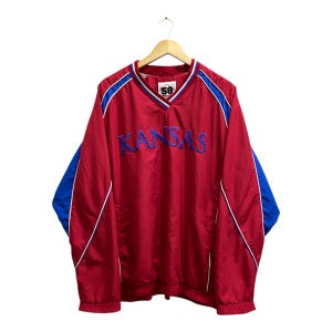 May include: A red and blue Kansas Jayhawks pullover jacket. The jacket has a V-neck, blue sleeves, and white trim. The word "KANSAS" is embroidered across the chest in blue. The number "58" is visible on the tag.
