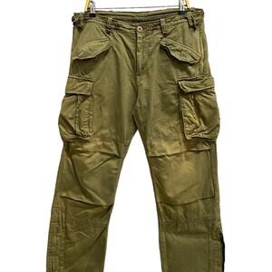 Puede incluir: Pantalones cargo de color verde oliva con cierre de botones, dos bolsillos delanteros, dos bolsillos laterales grandes con solapas abotonadas y dos bolsillos más pequeños encima. Las perneras incluyen cremalleras verticales en los laterales.