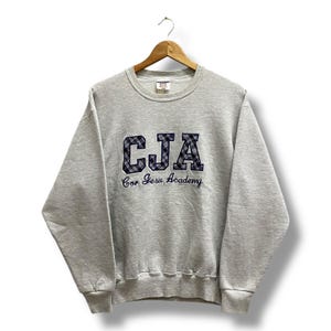 May include: A heather gray sweatshirt with the embroidered letters "CJA" in a plaid pattern, above the words "Cor Jesu Academy" in navy blue. The sweatshirt has a crew neck and long sleeves, and is hanging on a wooden hanger.