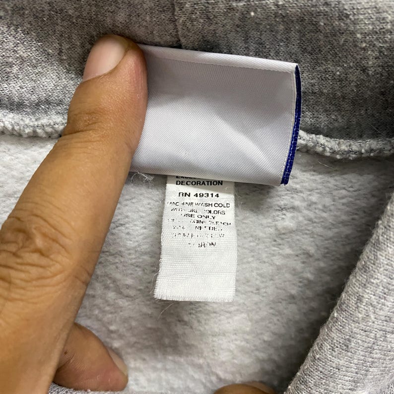 May include: Close-up of a gray hoodie with a white tag attached. The tag has the word "DECORATION" at the top, followed by the RN 49314 number. The tag also includes washing instructions. The hoodie is made of a soft, textured material.