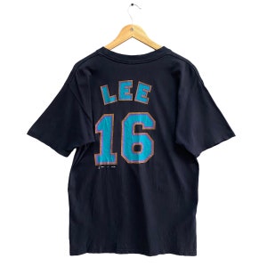 May include: Black t-shirt with the name "LEE" and the number "16" in teal and gold lettering on the back. The shirt is hanging on a wooden hanger against a white background. The shirt has short sleeves.