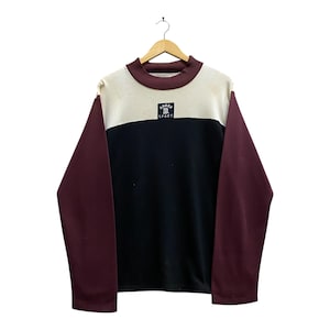 Peut inclure: Sweat-shirt à blocs de couleurs avec des panneaux crème, noirs et bordeaux. Le haut à manches longues présente un col montant et un petit écusson avec l'inscription "BANKS SPORT". Le sweat-shirt est suspendu à un cintre en bois.