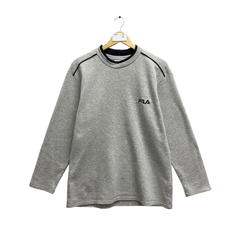 Puede incluir: Una sudadera gris de manga larga con el nombre de la marca "FILA" bordado en rojo en el pecho. La sudadera tiene un cuello negro y ribetes negros a lo largo de los hombros. La prenda est&aacute; colgada de una percha de madera.