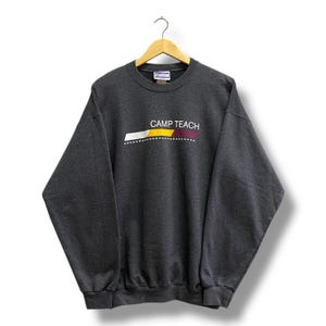 Op de afbeelding: Donkergrijze sweatshirt met de tekst "CAMP TEACH" in wit, geel en bordeauxrood. De sweatshirt hangt aan een houten hanger tegen een witte achtergrond. Het heeft lange mouwen en een ronde hals.