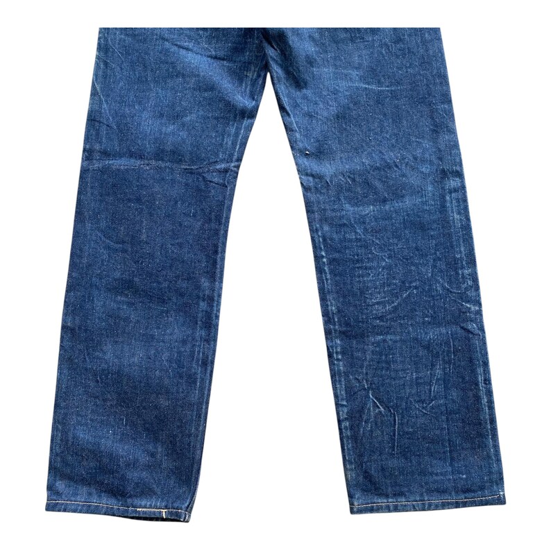 May include: A pair of dark blue denim jeans featuring a straight-leg cut with visible fading and whiskering patterns throughout the fabric. The denim has a textured appearance with subtle creasing and a classic stitched hem at the bottom of each leg.