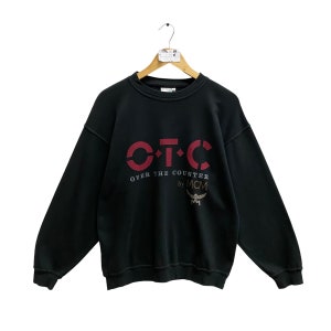 Puede incluir: Sudadera negra con el texto "O.T.C. Over the Counter by MCM" en color burdeos. La sudadera está colgada de una percha de madera. El logotipo de MCM es visible debajo.