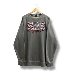 Könnte beinhalten: Graues Sweatshirt mit den Buchstaben "HHS" in einem Zickzackmuster und dem Wort "Tigers" in Burgunderrot. Das Sweatshirt hängt an einem Holzbügel vor weißem Hintergrund.