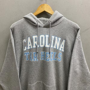 May include: A gray hooded sweatshirt with the text "CAROLINA TAR HEELS" in white and light blue lettering. The sweatshirt has a drawstring hood and a front pocket. The garment is hanging on a wooden hanger.