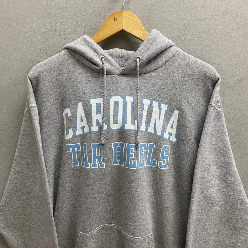 May include: A gray hooded sweatshirt with the text "CAROLINA TAR HEELS" in white and light blue lettering. The sweatshirt has a drawstring hood and a front pocket. The garment is hanging on a wooden hanger.