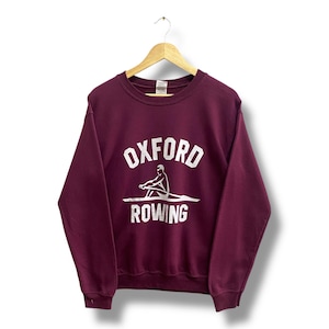Puede incluir: Una sudadera burdeos con las palabras "OXFORD ROWING" en letras blancas y un gráfico blanco de un remero. La sudadera está colgada en una percha de madera sobre un fondo blanco.