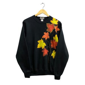 Può includere: Felpa nera girocollo con un design di foglie colorate. Le foglie sono nelle tonalità del giallo, arancione e rosso, con contorni dorati. La felpa è appesa a una gruccia di legno.