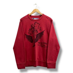 Peut inclure: Un sweat-shirt rouge à manches longues et col rond avec un motif noir représentant un arbre ailé. Le texte "Do not follow where the path may lead, go instead where there is no path and leave a trail" est imprimé en noir sur le côté droit.