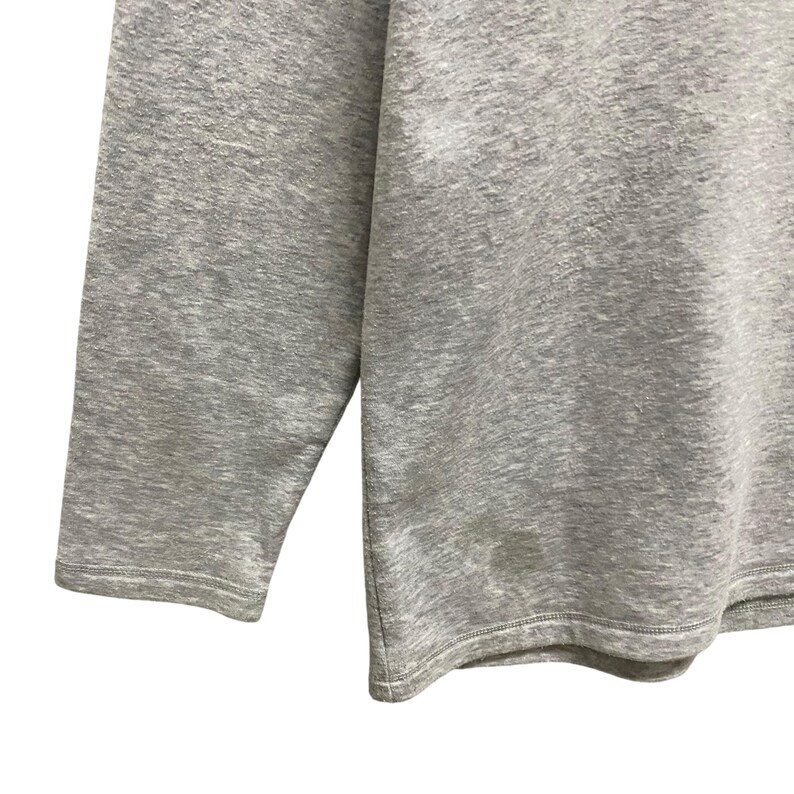 Puede incluir: Primer plano de una camiseta de manga larga gris jaspeado. La tela tiene un aspecto texturizado. La manga y el dobladillo son visibles, con una costura. La camiseta parece estar hecha de un material suave y c&oacute;modo.