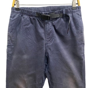 May include: Dark blue pants featuring an elastic waistband with an integrated black nylon webbing belt and a plastic side-release buckle. The pants have a standard fly construction, side pockets, and a straight-leg cut with visible fabric texture.