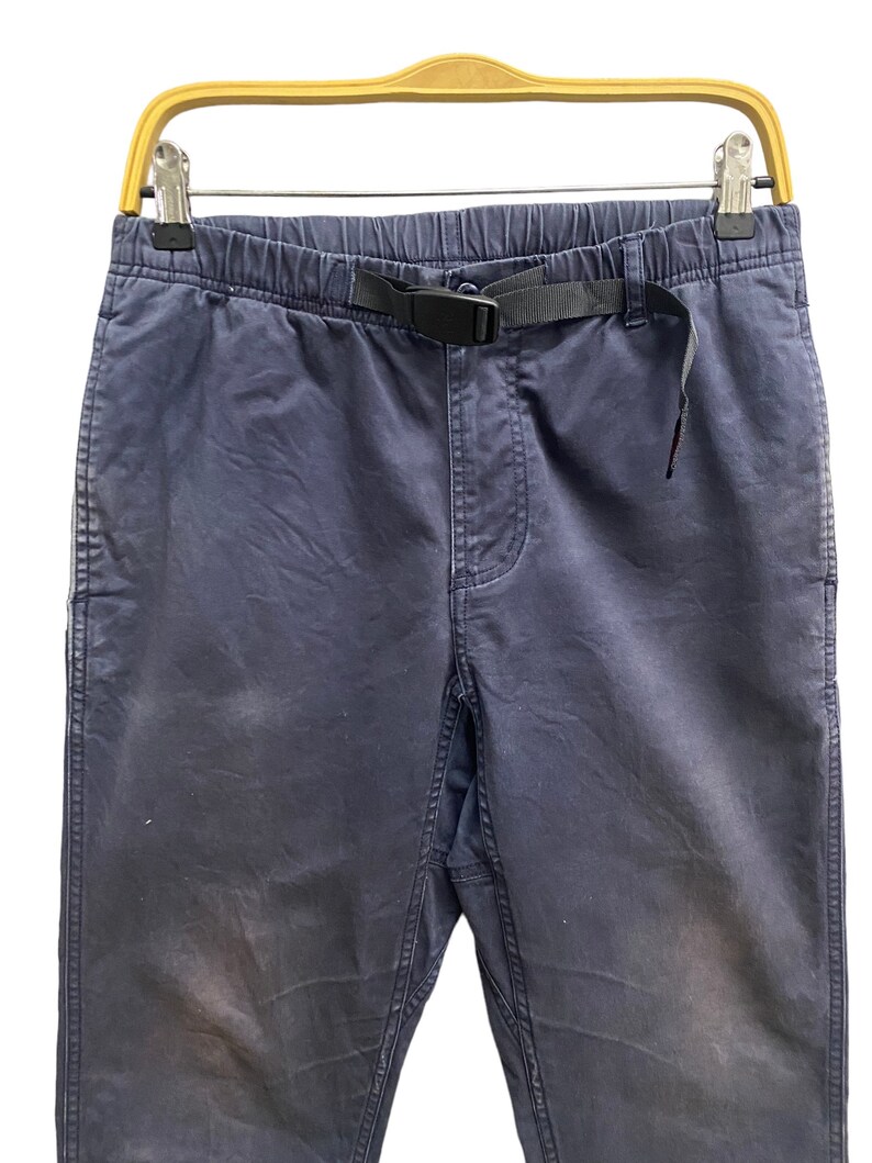 May include: Dark blue pants featuring an elastic waistband with an integrated black nylon webbing belt and a plastic side-release buckle. The pants have a standard fly construction, side pockets, and a straight-leg cut with visible fabric texture.