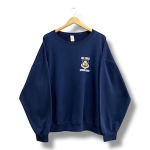 May include: Navy blue sweatshirt with the words "AIR FORCE" and "JUNIOR ROTC" printed on the front. The sweatshirt has a crew neck and long sleeves. The graphic is a crest with a gold and white design.