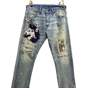 May include: Light blue denim jeans with distressed details, featuring a purple and white unicorn patch on the left leg and a black and white patch on the right leg.