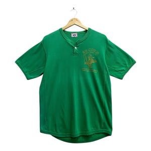 May include: Green short-sleeved Henley shirt with a three-button placket and a crew neck. The shirt has a gold graphic on the chest with the text "Sea Corn" and "American Favorite Heritage Fashion". It is hanging on a wooden hanger.