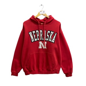 Puede incluir: Sudadera roja con capucha con la palabra "NEBRASKA" en letras blancas y negras y una "N" blanca en el centro.