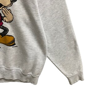 May include: A light gray sweatshirt featuring a graphic of Mickey Mouse in a classic pose. The cartoon character is depicted in black, red, yellow, and tan. The Disney logo is visible near the bottom of the graphic.