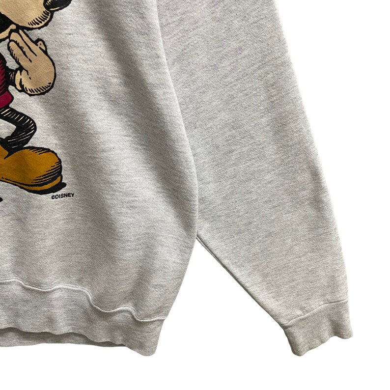 May include: A light gray sweatshirt featuring a graphic of Mickey Mouse in a classic pose. The cartoon character is depicted in black, red, yellow, and tan. The Disney logo is visible near the bottom of the graphic.