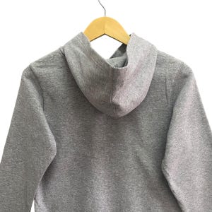 Puede incluir: Una sudadera con capucha gris jaspeado con capucha, colgada de una percha de madera. La sudadera con capucha est&aacute; hecha de un material suave y tiene un dise&ntilde;o simple y cl&aacute;sico. El fondo es blanco.