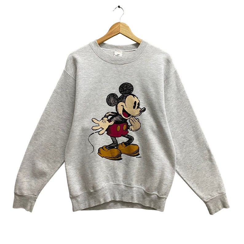 May include: A light gray sweatshirt featuring a classic Mickey Mouse graphic. The cartoon character is depicted in black, red, and yellow, with a smiling expression. The sweatshirt is hanging on a wooden hanger.