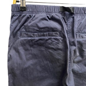 May include: A pair of dark navy blue pants featuring an elastic waistband with a black nylon webbing strap, a single horizontal welt pocket on the back, and visible stitching along the seams, suspended from a light-colored wooden hanger.