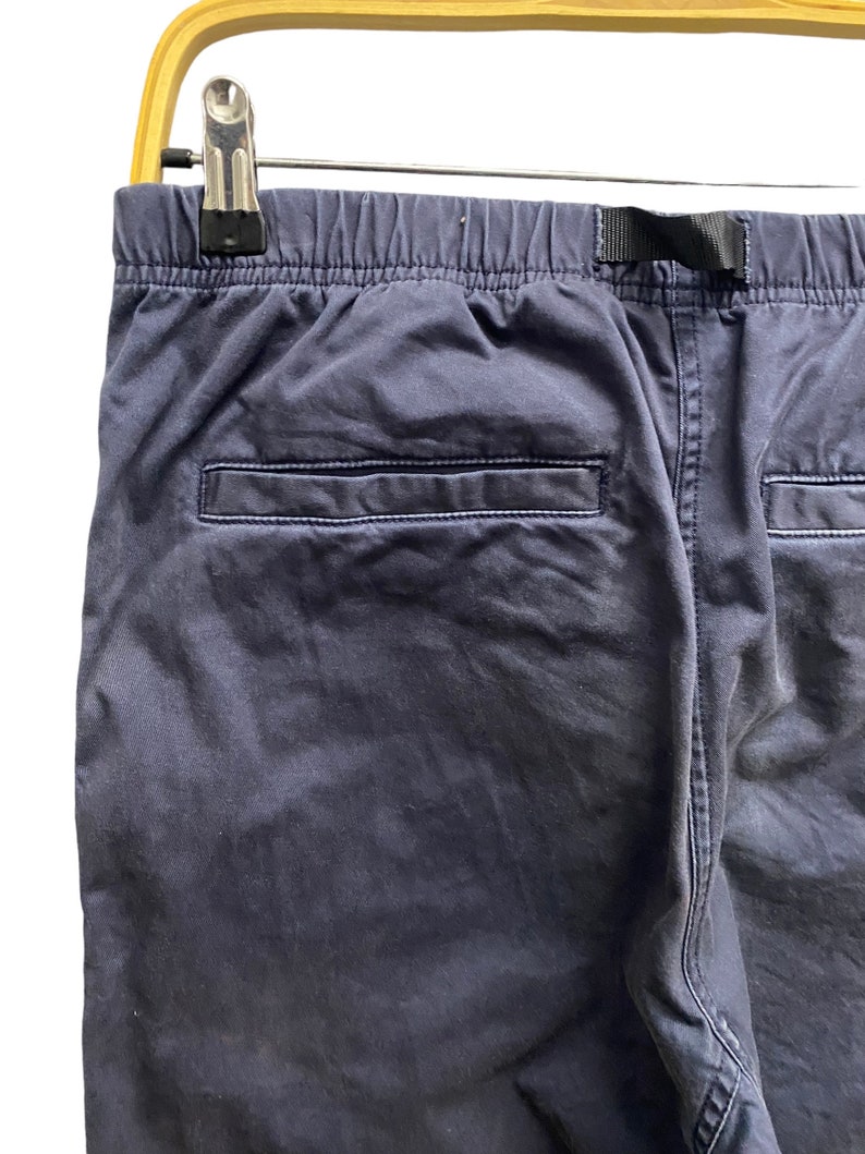 May include: A pair of dark navy blue pants featuring an elastic waistband with a black nylon webbing strap, a single horizontal welt pocket on the back, and visible stitching along the seams, suspended from a light-colored wooden hanger.