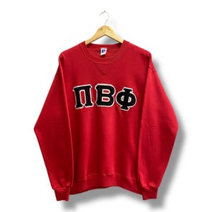 Könnte beinhalten: Ein rotes Sweatshirt mit den griechischen Buchstaben ΠΒΦ in Schwarz mit weißer Umrandung. Das Sweatshirt hat einen Rundhalsausschnitt und lange Ärmel. Das Russell Athletic-Logo ist sichtbar. Das Sweatshirt hängt an einem Holzbügel.