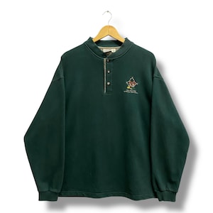 May include: Dark green long-sleeve sweatshirt with a button-up collar. The sweatshirt has a small embroidered design on the chest with text that reads "THE BAD DOG NATIONAL PARK". The sweatshirt is hanging on a wooden hanger.