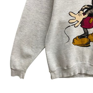 May include: A light gray sweatshirt featuring a graphic of Mickey Mouse. The cartoon character is depicted in black, red, yellow, and tan. The sweatshirt has a crew neck and long sleeves.