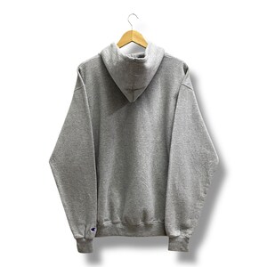 May include: A gray Champion hoodie, viewed from the back, hangs on a wooden hanger. The hoodie features a hood, long sleeves, and a ribbed hem and cuffs. The Champion logo is visible on the left cuff. The background is white.