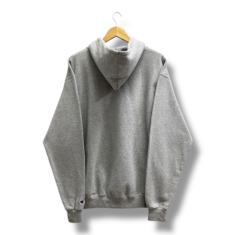 May include: A gray Champion hoodie, viewed from the back, hangs on a wooden hanger. The hoodie features a hood, long sleeves, and a ribbed hem and cuffs. The Champion logo is visible on the left cuff. The background is white.