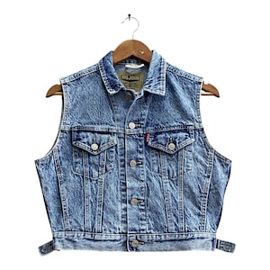 May include: A light blue denim vest with a button-up front, two chest pockets, and a small red tag. The vest is hanging on a wooden hanger. The label reads "BIG JOHN".