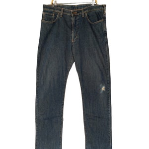 May include: A pair of dark blue denim jeans featuring a classic five-pocket design, a button fly closure, and a subtle distressed mark on the left thigh. The jeans are constructed with visible stitching and are displayed hanging on a wire hanger.