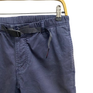May include: Navy blue trousers featuring an elasticized waistband and an integrated black nylon webbing belt with a plastic side-release buckle. The pants include a front fly detail and are suspended from a light-colored wooden clothes hanger.